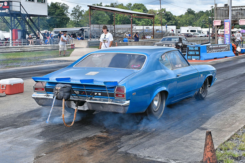 A bright blue classic car with a racing number is accelerating down a drag strip, producing smoke from its tires. In the background, spectators watch from the sidelines, and the racing track is clearly visible.