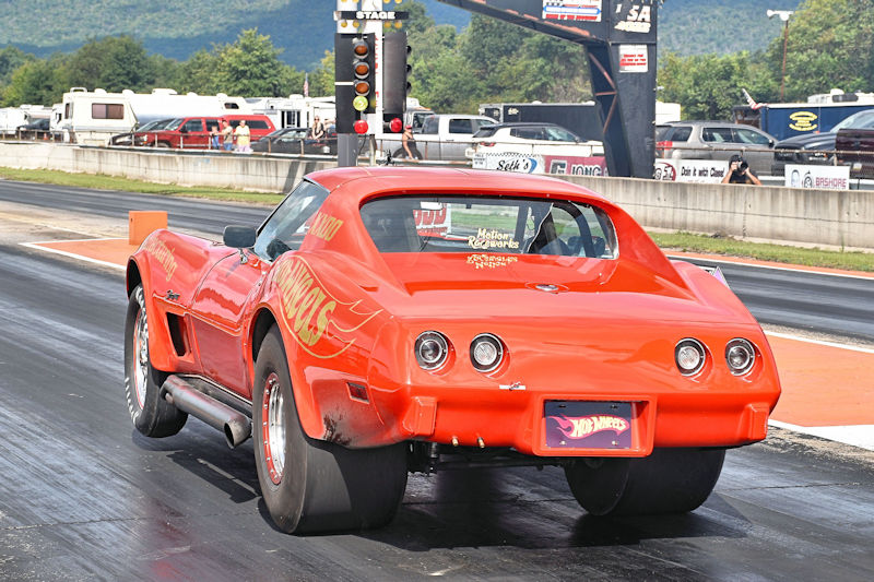 A classic orange drag racing car on a racetrack, featuring a unique flame design and large rear tires. The car is positioned at the starting line, with racing lights in the background.