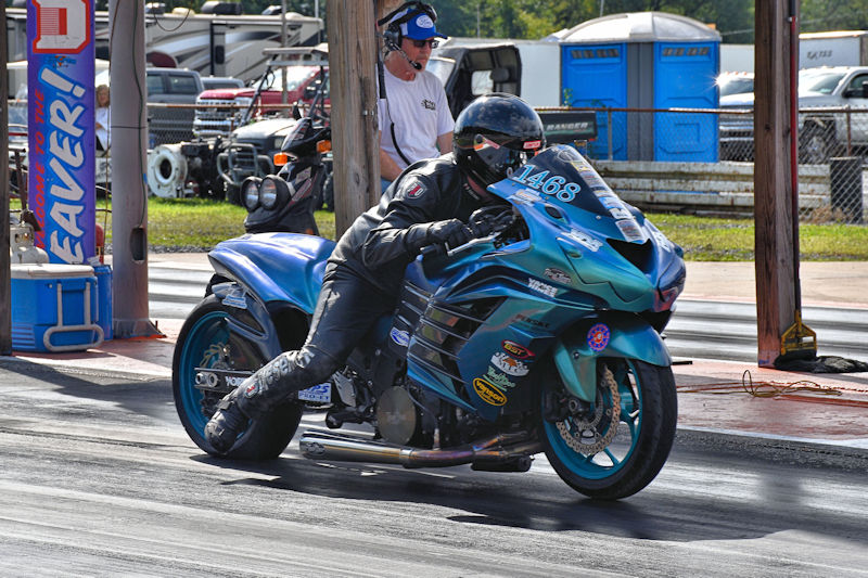 A racer in a black leather suit riding a blue and turquoise motorcycle on a drag racing track, with a spectator in the background.