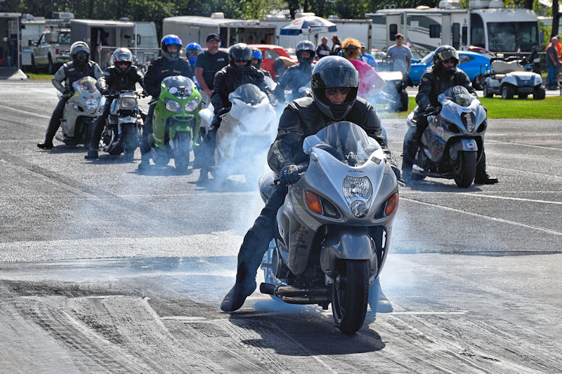 Motorcycle riders in helmets and leather jackets preparing to race, with smoke trailing from the front motorcycle on a racetrack.
