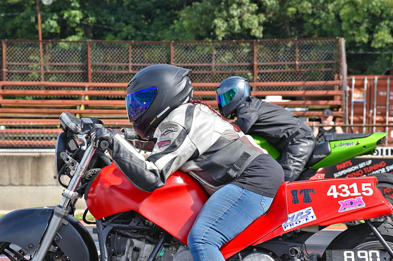 Two motorcycle racers in a starting position on a drag strip, one on a red bike and the other on a green bike, both wearing helmets and racing gear.