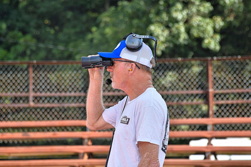 A man wearing a baseball cap and headphones uses binoculars while standing near empty bleachers in a natural setting.