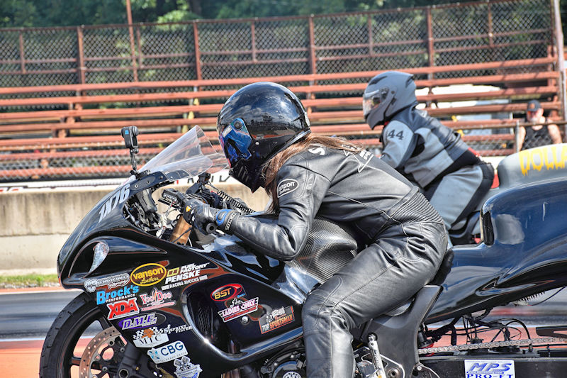 A motorcycle racer in black leather gear prepares for a race on a drag strip, with another competitor in the background.