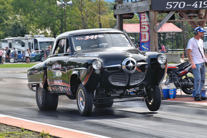 A black drag racing car performing a wheelie on a racetrack, with spectators in the background and a man standing nearby.