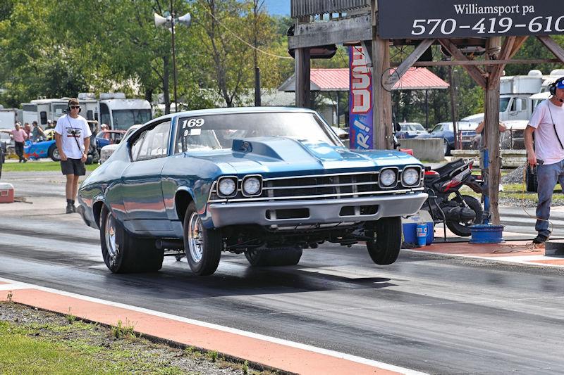 A blue classic car performing a wheelie on a drag strip, with spectators and a timing tower in the background.