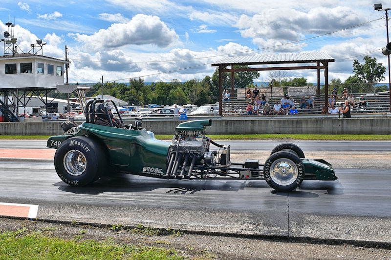 A green dragster racing down a track with spectators in the background and a cloudy sky.
