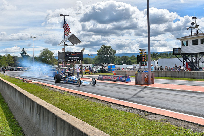 A drag racing vehicle speeding down the track with smoke trailing behind, under a bright blue sky with clouds.