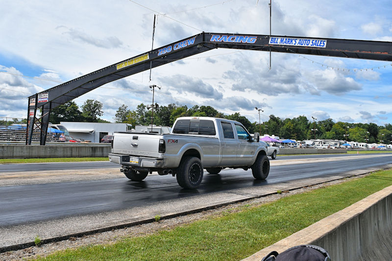 A silver pickup truck accelerating down a drag racing track under a banner for Bill Mark's Auto Sales.
