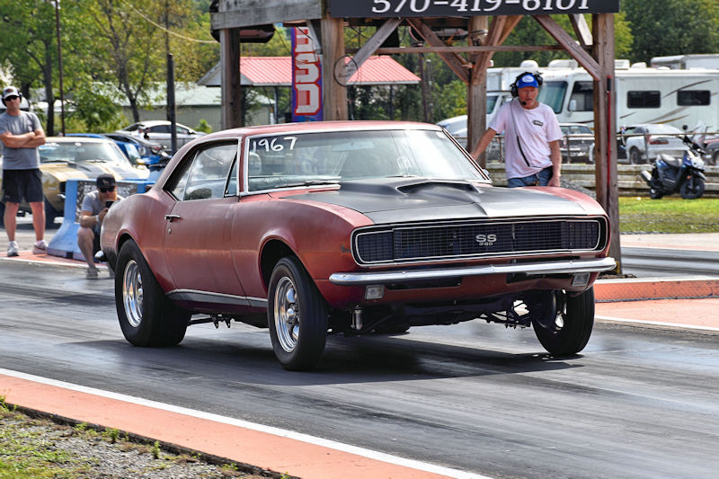 A classic 1967 Chevrolet Camaro SS racing down a drag strip, with spectators in the background.