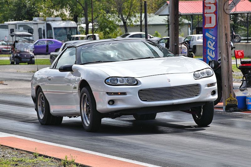 A white car performing a wheelie on a drag strip, with a blurred background of spectators and vehicles.