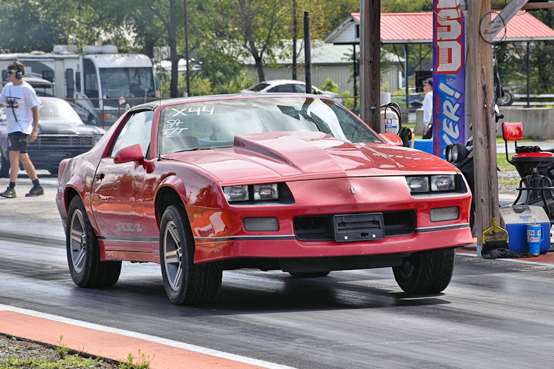 A red sports car, identified as a Camaro, is racing on a drag strip with its front tires lifted off the ground. A person is visible in the background, preparing for the next race.