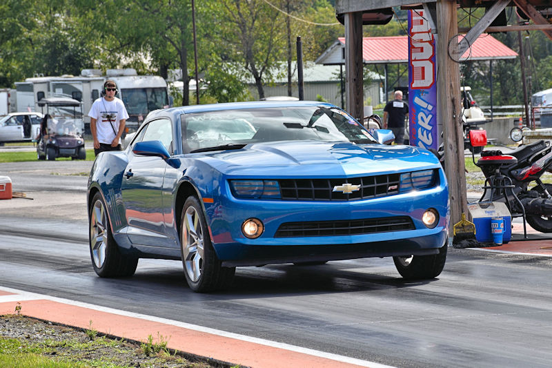 A blue Chevrolet Camaro approaching the starting line of a drag racing track, with a person in a white shirt in the background.