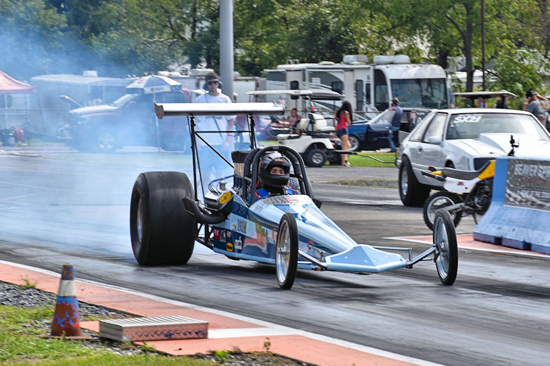 A drag racer in a blue race car accelerates down the track, emitting smoke from the rear tires, with spectators in the background and other vehicles parked nearby.