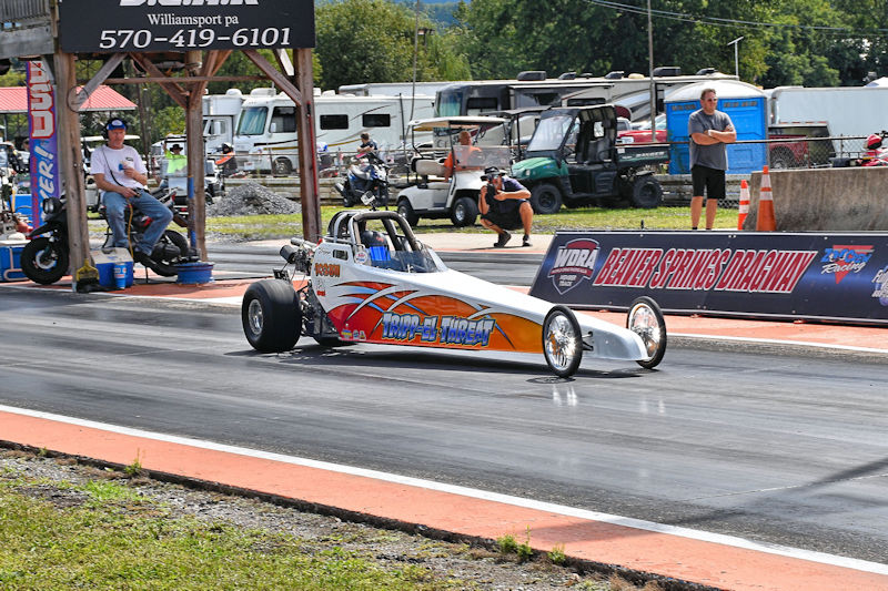 A drag racing car with colorful graphics moves down the track at Beaver Springs Dragway, with spectators watching and various vehicles parked in the background.