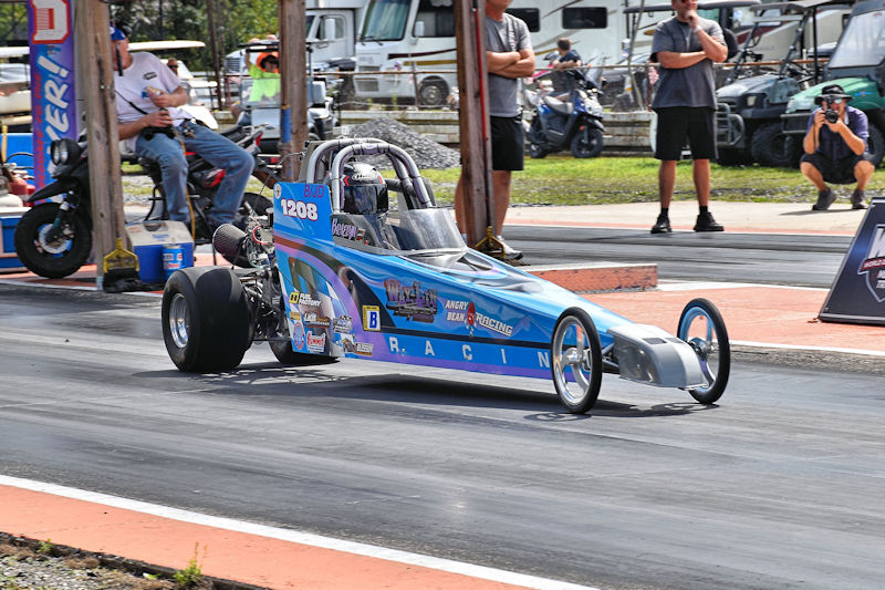 A drag racing car accelerating down the track with a driver in a helmet, spectators watching from the sidelines.