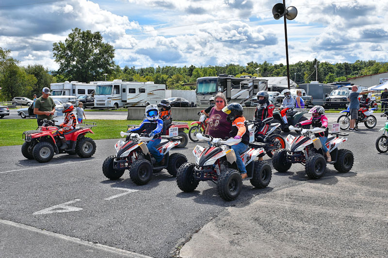 Group of children on ATVs lined up at a dirt track event with spectators and RVs in the background.