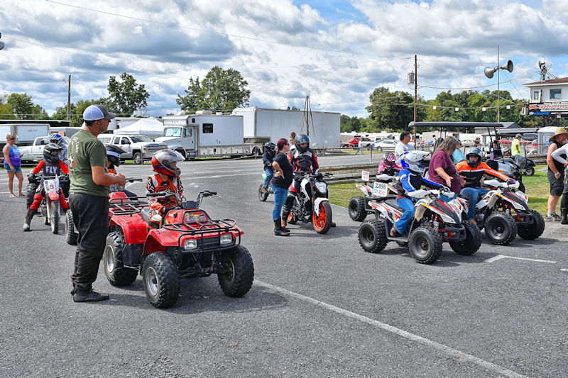 A group of people interacting around various ATVs and dirt bikes in a parking area, with participants wearing helmets and racing gear.