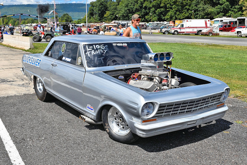 A silver drag racing car with the name 'QUICKSILVER' on the side, featuring a powerful engine and large rear tires, parked on a racetrack with spectators in the background.