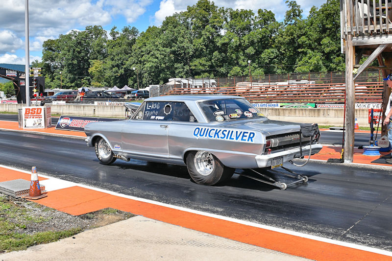 A silver racing car, labeled 'QUICKSILVER,' parked on a drag racing track under a partly cloudy sky, with spectators and a timing tower in the background.