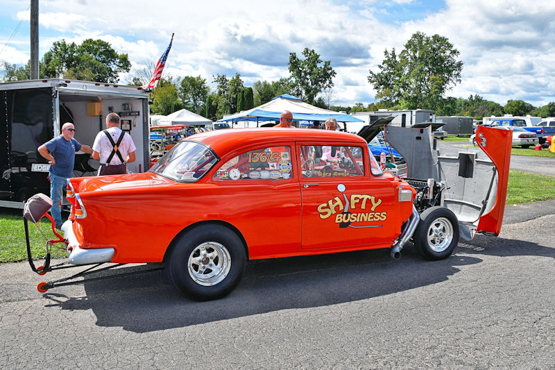 A vintage orange race car with the words 'SHI FY BUSINESS' on the side, parked with its hood open. Several people are gathered nearby, and there are tents and vehicles in the background under a partly cloudy sky.