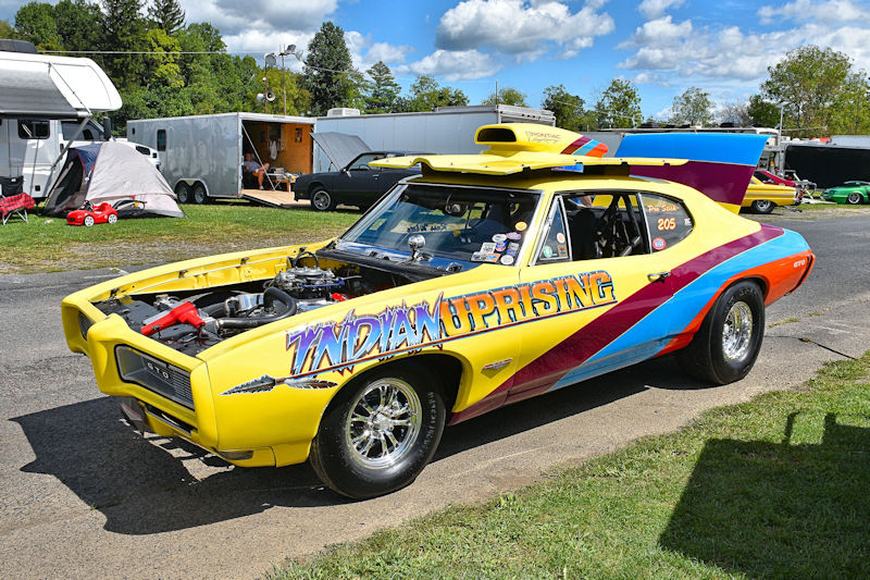 A bright yellow vintage race car with the name 'Indian Uprising' displayed on the side, parked on a street with trailers and other vehicles in the background.