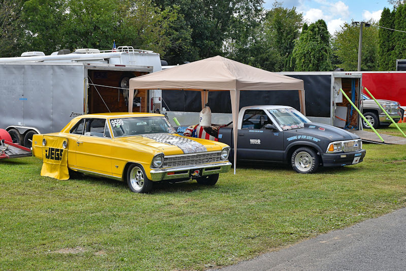 A bright yellow classic car with racing decals parked next to a gray pickup truck under a tent, with a trailer in the background at a car show event.