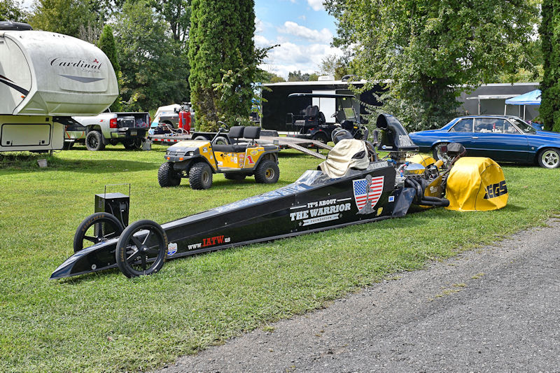 A drag racing car named 'The Warrior' on a grassy area, surrounded by various vehicles including an ATV and an RV. In the background, there are trees and a blue sky.