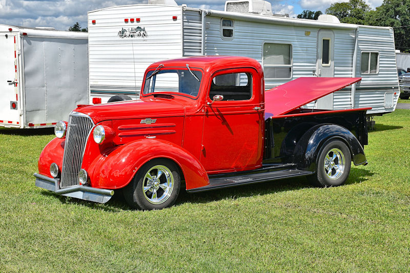 A vintage red pickup truck with a shiny chrome grille, parked on a grassy area, with a trailer in the background.