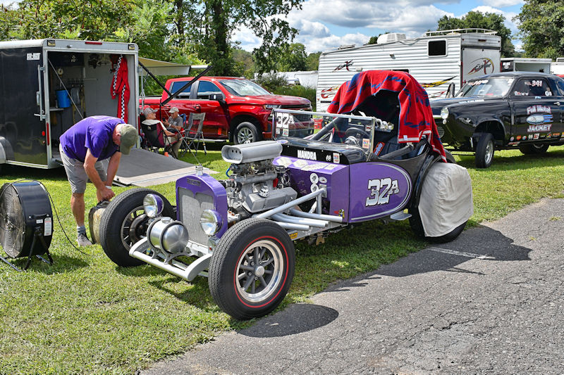 A vintage purple race car with an open engine and distinctive wheels is parked on a grassy area. A person in a purple shirt is adjusting a tire next to the car, while a trailer and other vehicles are visible in the background.