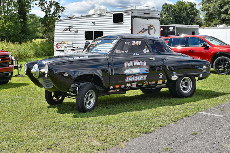 A black racing car with 'Full Metal Jacket' branding parked on grass, surrounded by trailers and other vehicles.