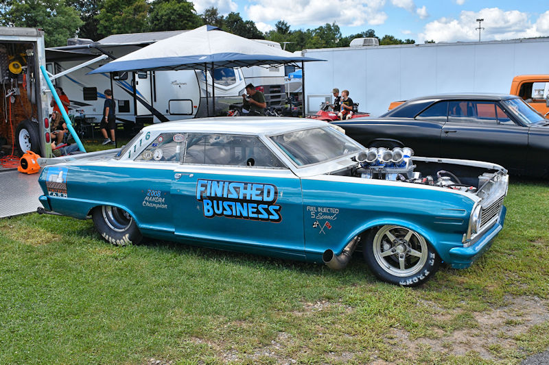 A classic blue racing car with the words 'FINISHED BUSINESS' printed on the side, parked on grass among other vehicles. The car features a distinctive hood scoop and chrome wheels.