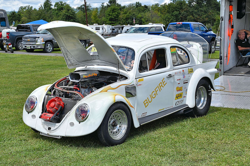 A modified white Volkswagen Beetle race car with an open hood, showcasing its engine. The car features racing graphics and is parked on grass, with other vehicles visible in the background.