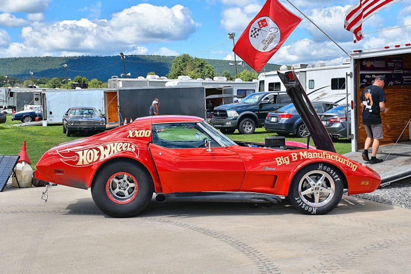 A red hot rod vehicle with the 'Hot Wheels' logo, parked with its hood open. It features racing tires and is surrounded by trailers and other cars in a grassy area, with clouds and hills in the background.