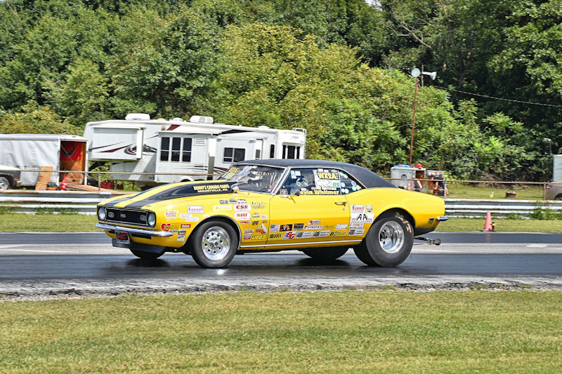 A yellow drag racing car accelerates down a track with various sponsor stickers visible on its body.