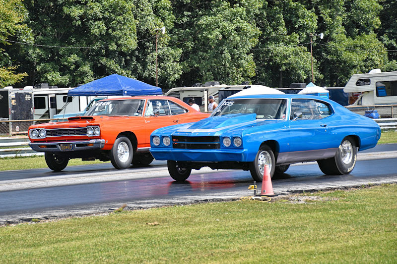 Two classic muscle cars racing down a drag strip, one orange and one blue, with a background of green trees and tents.