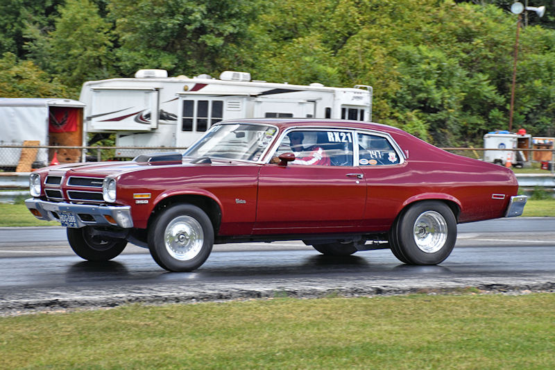 A vintage maroon muscle car racing down a drag strip, showcasing its chrome wheels and sleek design, with trailers in the background.