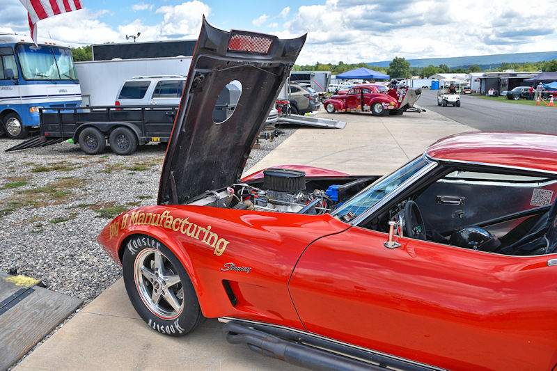 A red sports car with the hood raised, displaying the engine. The car is parked in a racing area, surrounded by trailers and other vehicles.