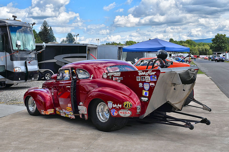 A classic red racing car with stickers parked in a paddock area, alongside an orange vehicle and camping trailers under a blue sky with clouds.