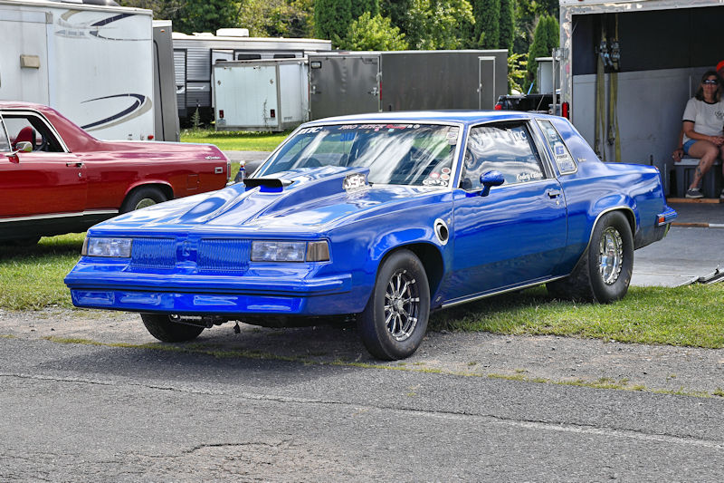 A vibrant blue racing car parked on the side of a track, with a sleek design and shiny black wheels, alongside another red car in the background.