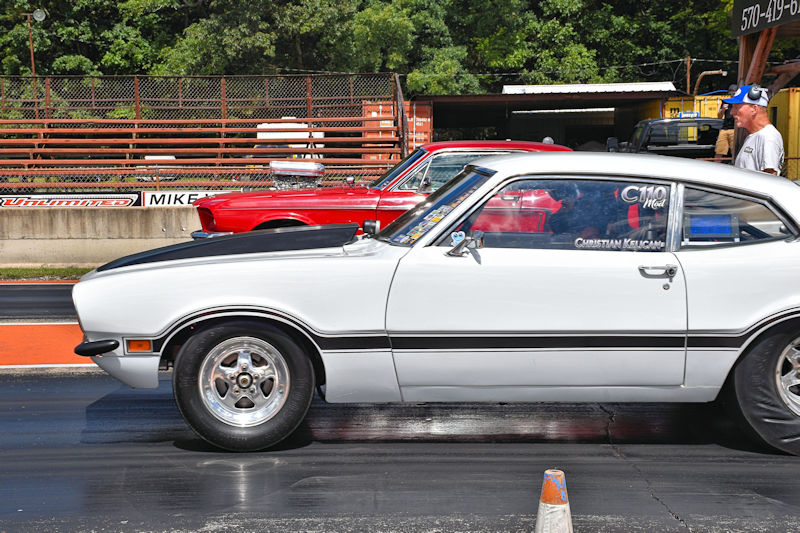 A white muscle car with a black stripe is positioned on a drag strip, ready for a race against a red car in the background. A spectator is visible in the background.