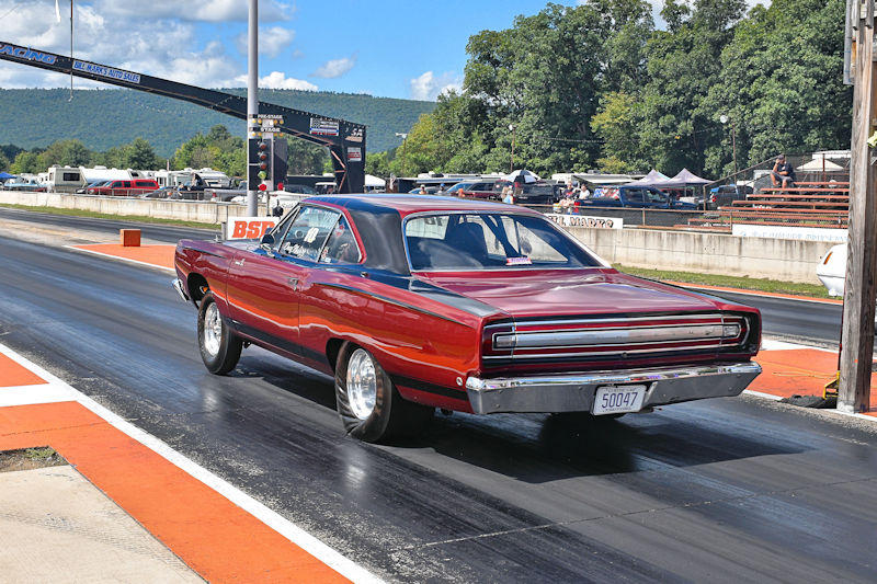 A classic red muscle car with a black roof is driving down a drag racing track, with spectators and trees in the background under a partly cloudy sky.