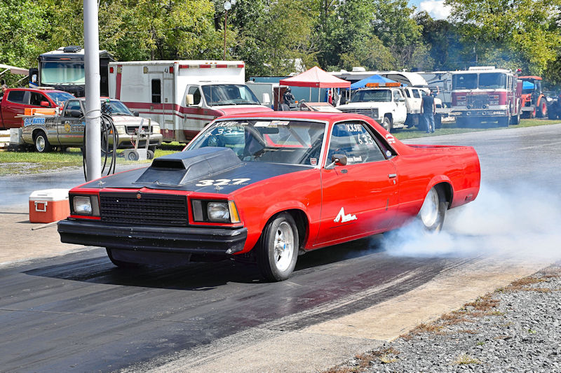 A red car performing a burnout at a drag racing event, with smoke coming from the rear tires and spectators in the background.