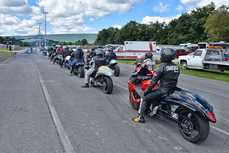 A group of motorcycle racers lined up on a track, preparing for a race, with some smoke visible in the distance and vehicles parked nearby.