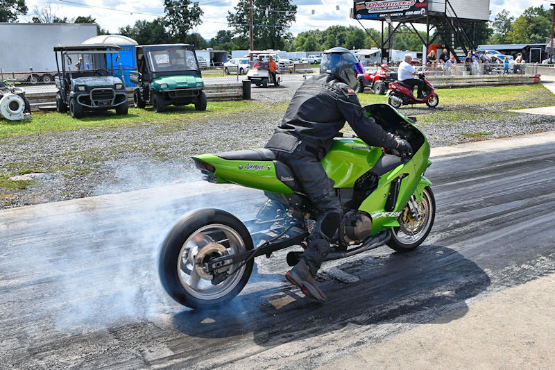 A motorcyclist in a black leather suit revving a green sports bike on a drag strip, creating tire smoke, with spectators and vehicles in the background.