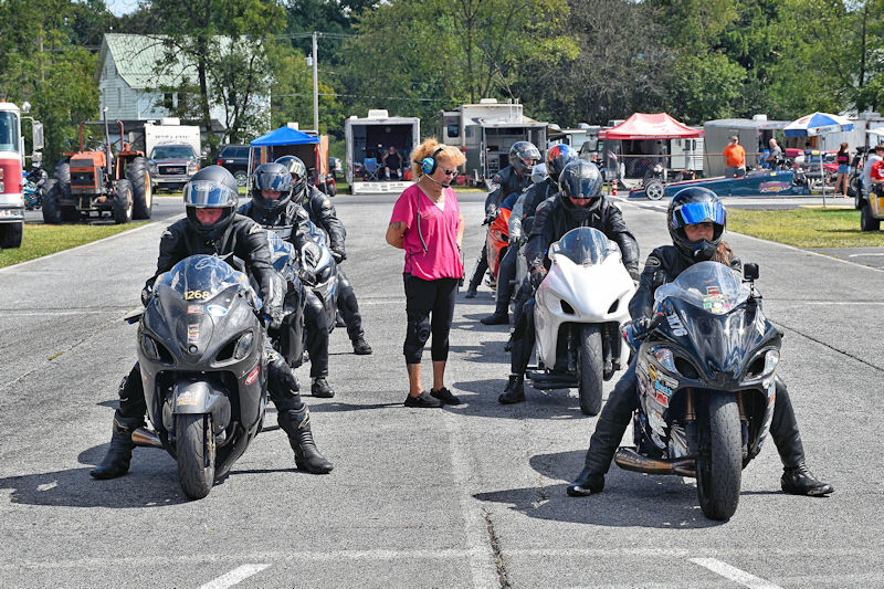 A group of motorcycle racers in gear, lined up on a track, with a woman in a pink shirt standing in front holding a clipboard. Various service vehicles and tents are visible in the background.