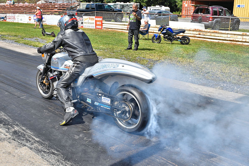 A motorcycle racer in leather gear performing a burnout on a drag strip, with smoke billowing from the rear tire. Spectators are seen in the background, and a smaller motorcycle is parked nearby.