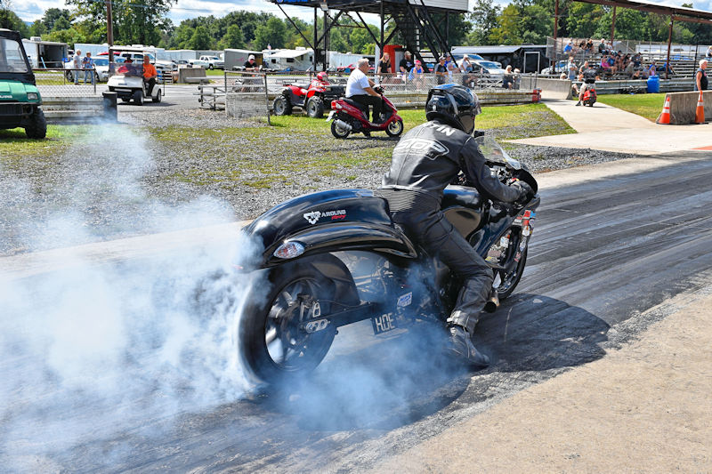 A motorcycle performing a burnout on a drag strip, with smoke rising from the tires. The rider is dressed in black and the scene is set during a sunny day at a race event.