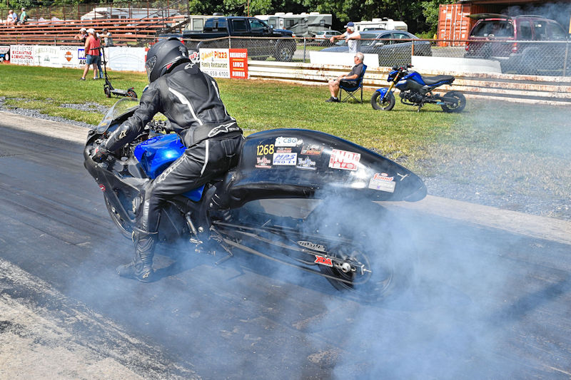 A motorcycle racer in a black leather suit performing a burnout, creating smoke on a drag strip. The scene features spectators in the background and a blue motorcycle parked nearby.