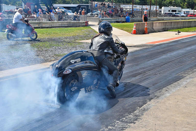 A motorcyclist in a black racing suit is accelerating on a dragstrip, producing smoke from the rear tire. In the background, spectators are watching from the stands, while another vehicle is visible nearby.