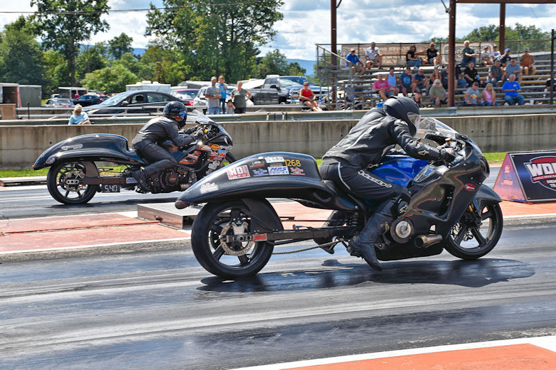 Two motorcycles racing on a drag strip, with riders in black leather suits, during a sunny day. Spectators are visible in the background.